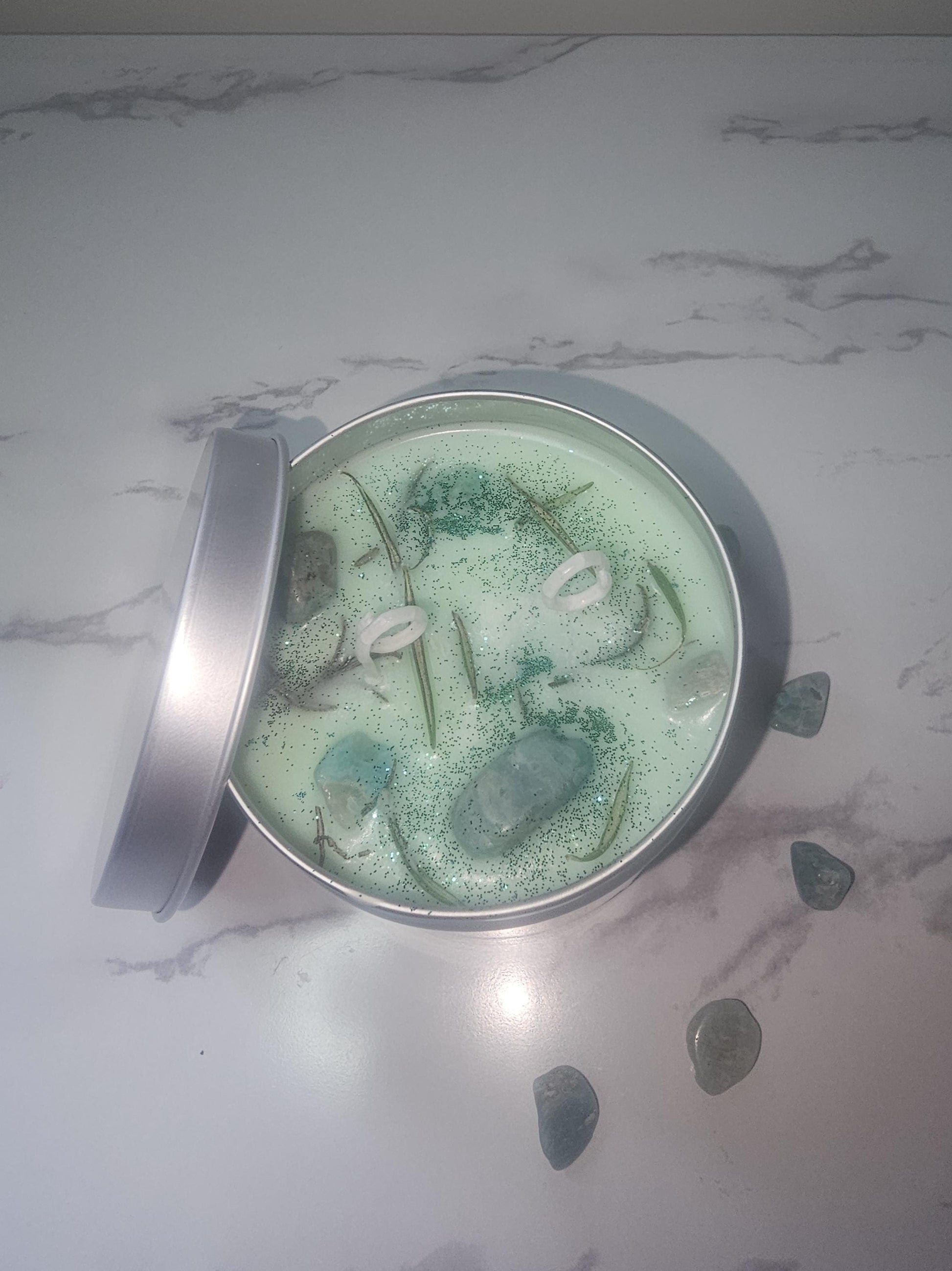 An above sot of a silver candle tin filled with light green scented soy wax and topped with green glitter, dried rosemary and amazonite crystals.