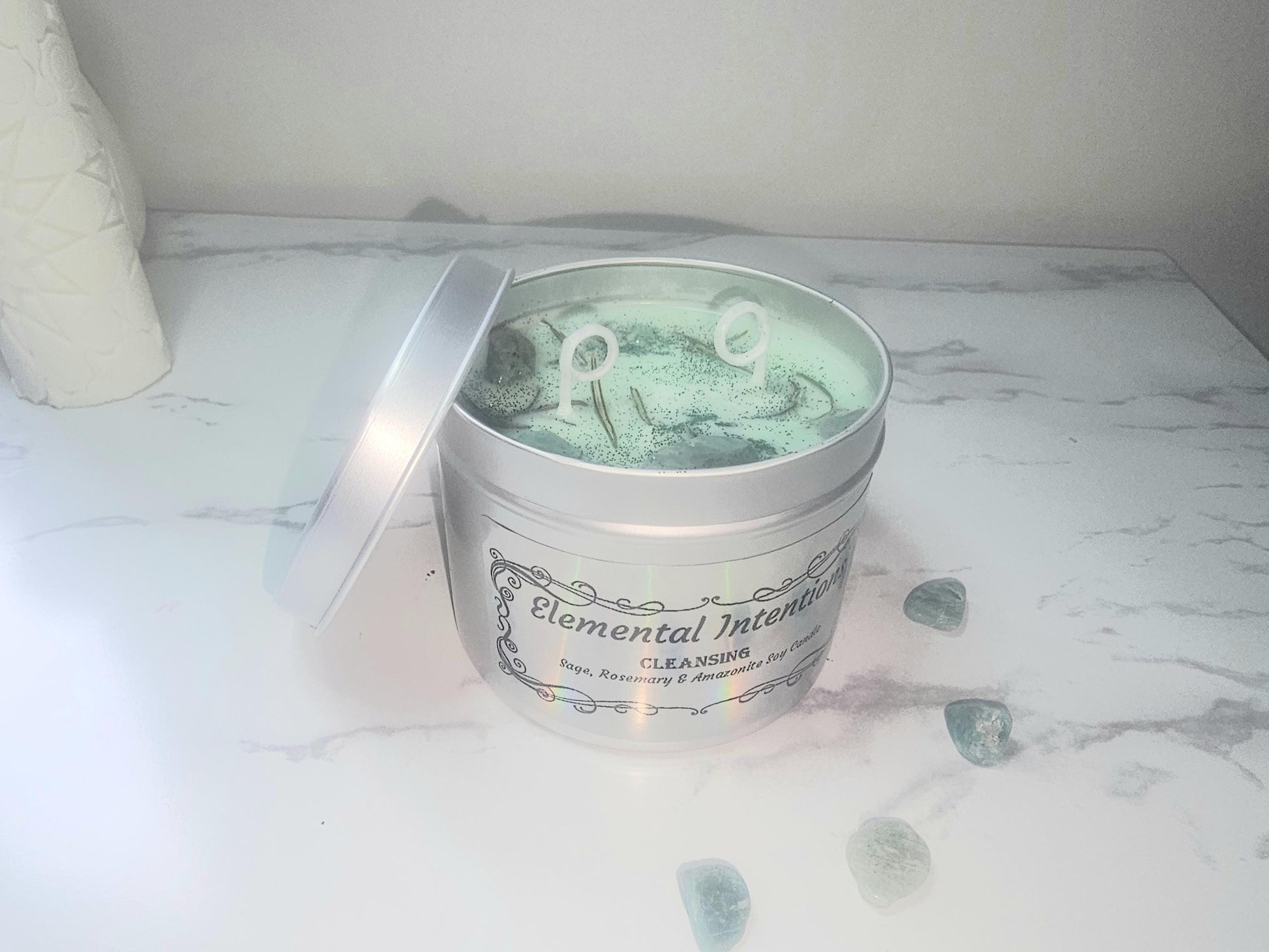 A silver candle tin filled with green scented soy wax and topped with dried rosemary and amazonite crystals There is a holographic label on the front that says &#39;elemental intentions&#39;, &#39;cleansing&#39; and &#39;sage, rosemary and amazonite soy candle&#39;.