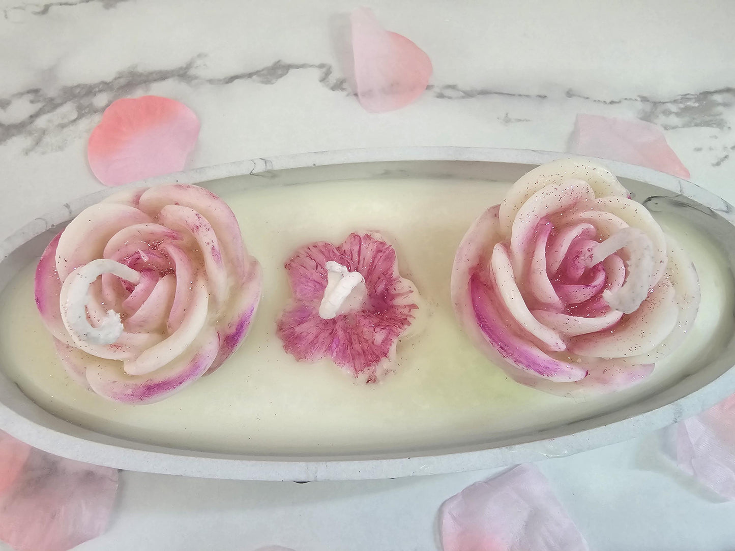 An above shot of an oval marble concrete resin candle holder filled with white sox wax and topped with two ornate wax white and pink roses and a pink lotus flower in the middle, surrounded by rose petals.