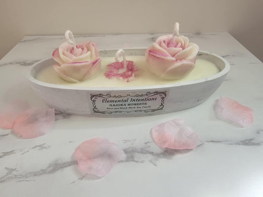 An oval hand poured concrete resin marble candle holder filled with white sox wax and topped with 2 ornate rose wax candles and a smaller lotus candle in the middle. The label says &#39;elemental intentions&#39; &#39;garden moments&#39; and &#39;rose and black musk soy candle&#39;.