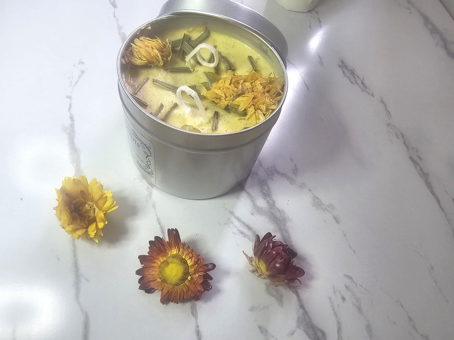 An above shot of a yellow candle in a silver container on a white marble background. The candle is surrounded by yellow and red decorative flowers and topped with lemongrass pieces, calendula lowers and citrine crystals, finished with a sprinkle of silver glitter.