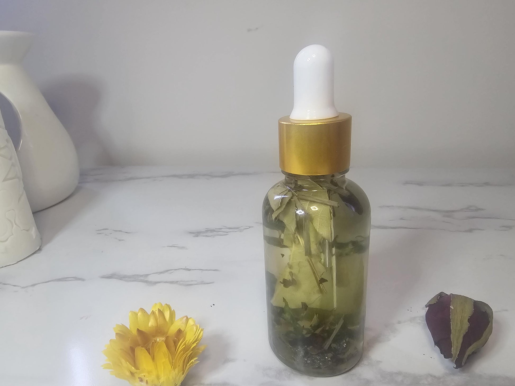 A clear dropper bottle with a gold top on a white marble background surrounded by red and yellow flowers. The bottle is filled with clear oil and different green herbs.