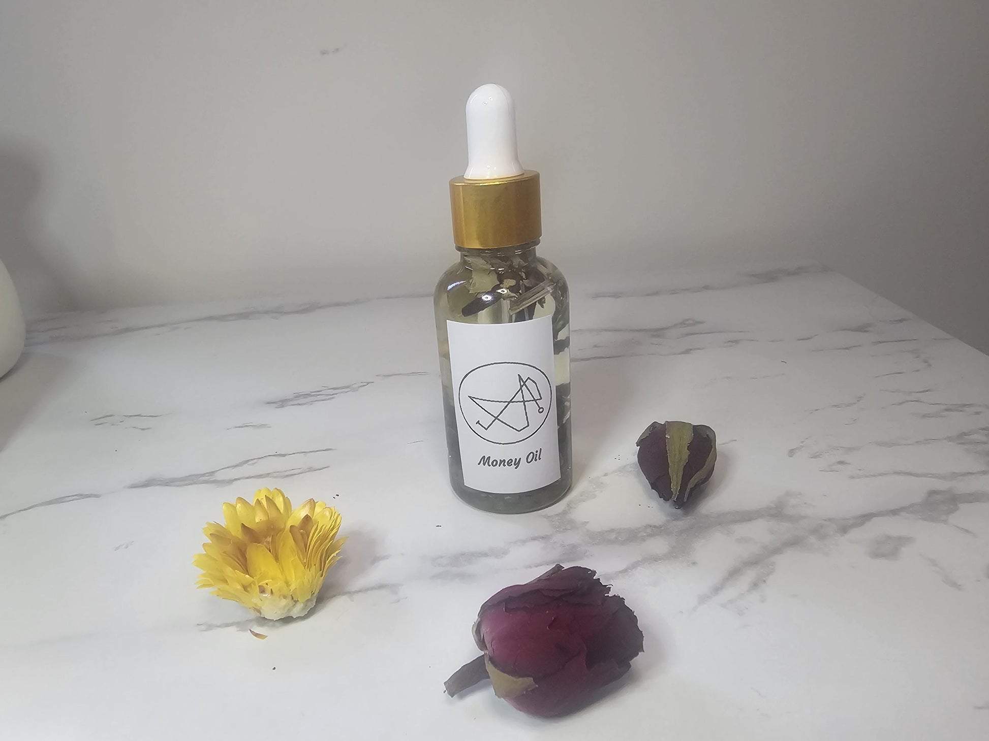 A clear dropper bottle with a gold top on a white marble background and surrounded by flowers. The front label contains a picture sigil and the words &#39;money oil&#39;.