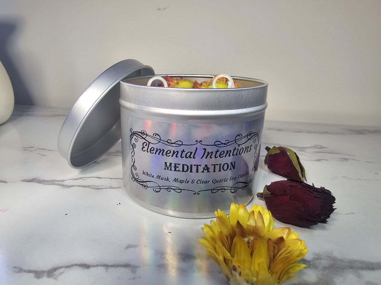 A front shot of a silver candle tin with a holographic label filled with green scented soy wax and topped with chrysanthemums and clear quartz crystals.