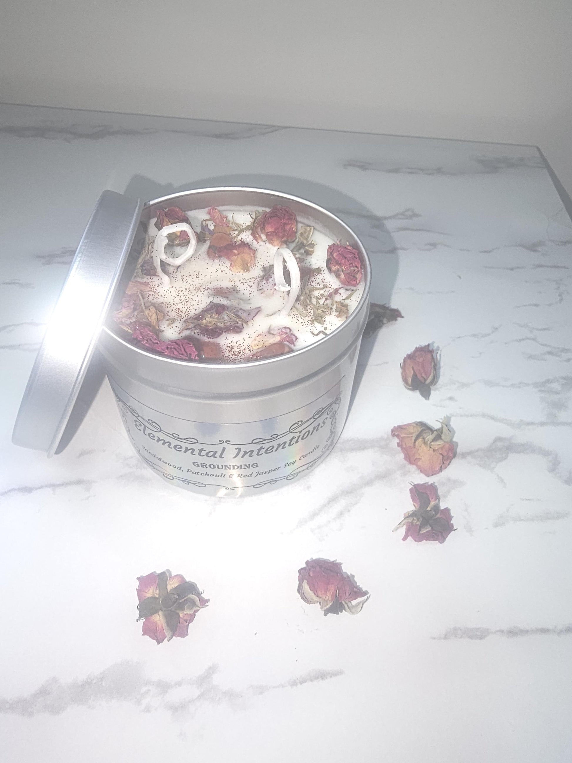 A silver candle tin filled with a nude coloured wax and topped with red jasper crystals and red flowers and glitter. The label on the front is holographic and says &#39;elemental intentions&#39;, &#39;grounding&#39; and &#39;sandalwood, patchouli and red jasper candle&#39;.