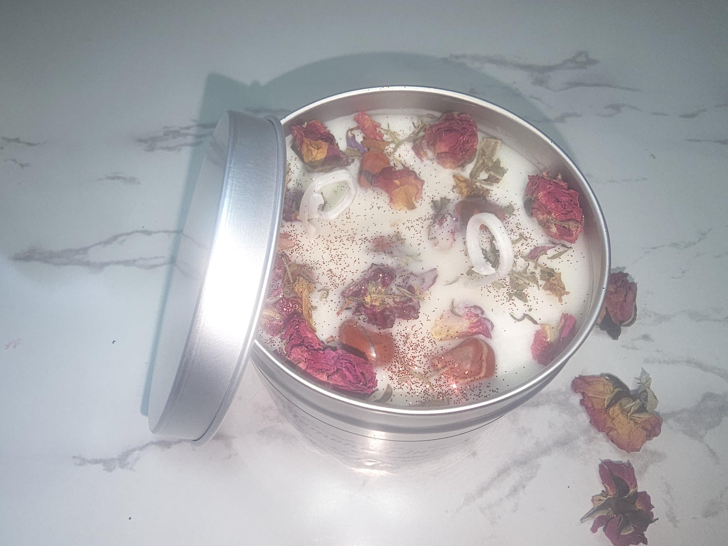 A silver candle tin on a white background surrounded by rose buds. The tin is filled with a nude cream coloured scented soy wax and topped with red jasper crystals, a sprinkling of red glitter and red rose buds.