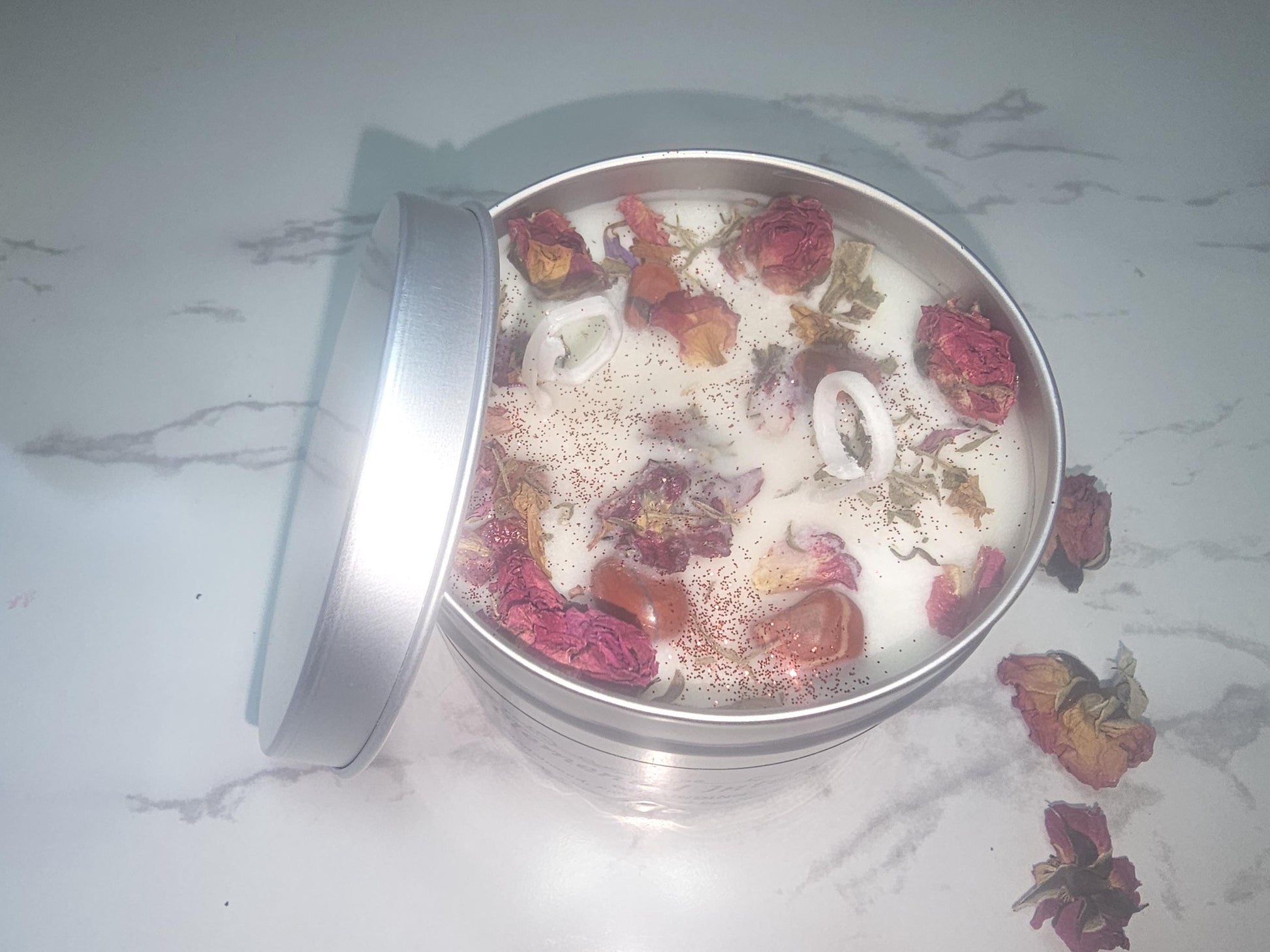 A silver candle tin on a white background surrounded by rose buds. The tin is filled with a nude cream coloured scented soy wax and topped with red jasper crystals, a sprinkling of red glitter and red rose buds.